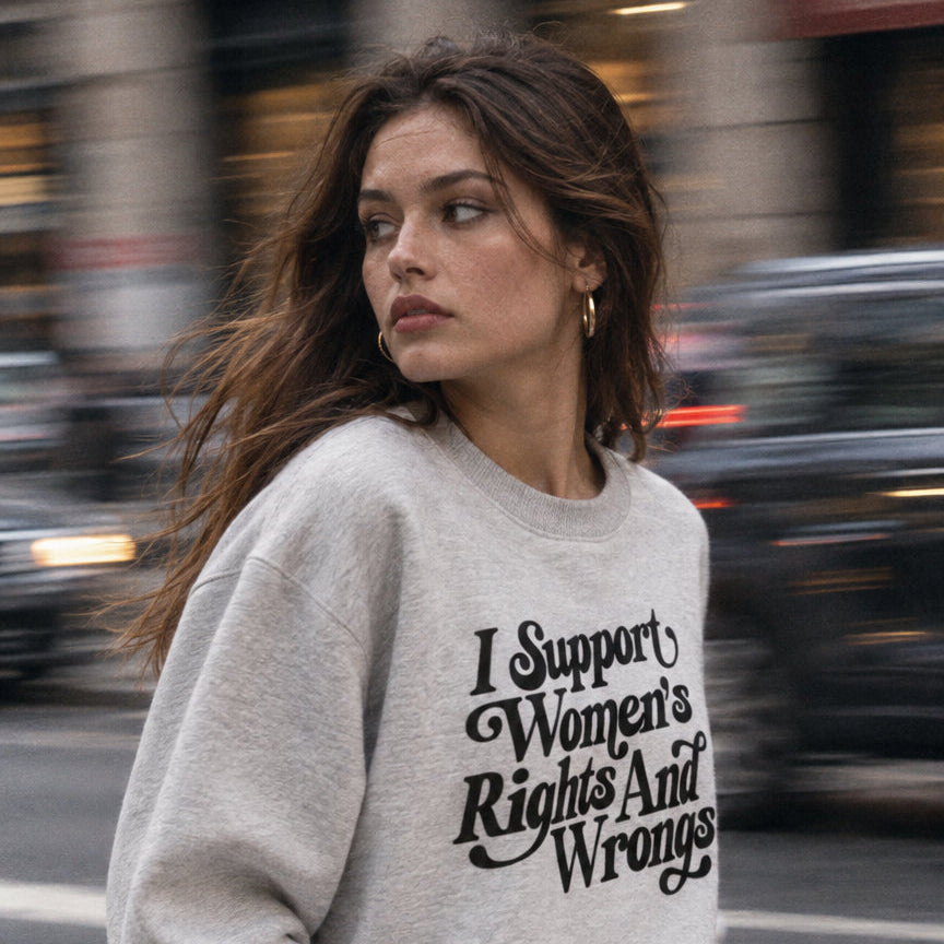 A woman walking on a city crosswalk wearing a light gray sweatshirt with the text 'I Support Women's Rights And Wrongs' in black script, paired with blue jeans.