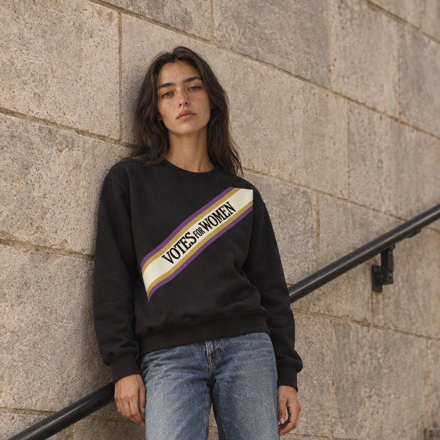 A woman stands on stone stairs wearing a black crewneck sweatshirt with a diagonal 'VOTES WOMEN' graphic in yellow, white, and purple, paired with light-wash blue jeans and white sneakers.