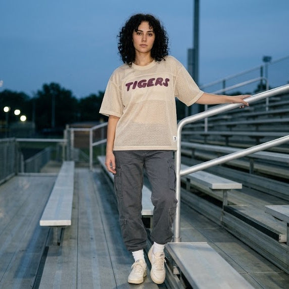 A model stands on the walkway between bleachers at a sports field during dusk, wearing a beige graphic t-shirt with 'TIGERS' printed in dark maroon across the chest, dark gray sweatpants, and white sneakers.