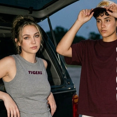 Three models wearing streetwear apparel posing in a parking lot at dusk. The central male model wears a maroon t-shirt with 'TIGERS' printed vertically in white, paired with off-white cargo pants.