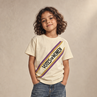 A young girl with curly brown hair stands in a studio wearing a cream-colored t-shirt with a diagonal purple and gold stripe that reads 'VOTES FOR WOMEN', paired with blue jeans and white sneakers.
