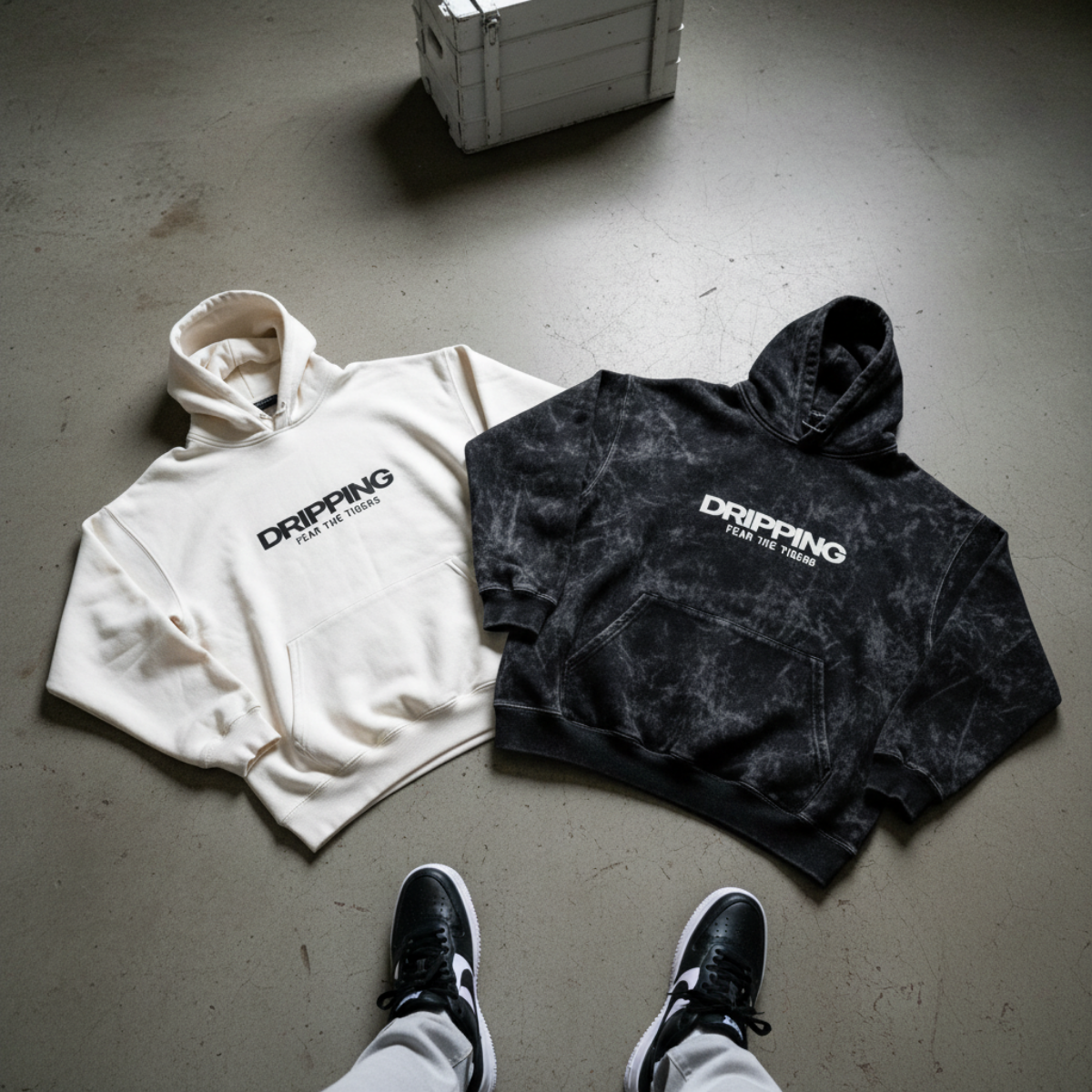 Two oversized streetwear hoodies, one cream and one black tie-dye, with 'DRIPPING FEAR THE TIGERS' text, laid on an urban floor beside sporty sneakers.