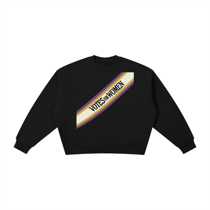 A black long-sleeve crewneck sweatshirt with a diagonal graphic on the chest featuring a cream, purple, and gold stripe with the text 'VOTES FOR WOMEN' in black.