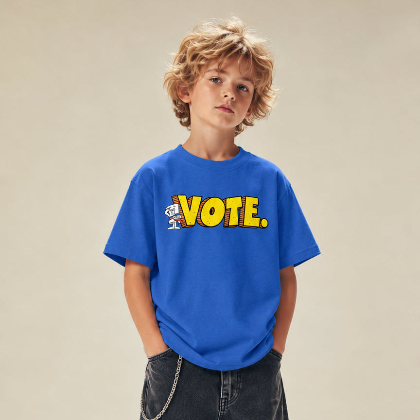 A young boy with curly blond hair stands against a warm beige studio background wearing a bright blue graphic t-shirt with yellow 'VOTE.' lettering, dark gray baggy jeans, and black sneakers.