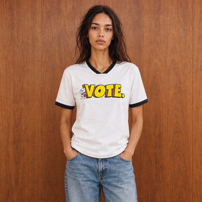 A woman stands against a wood-paneled wall wearing a white ringer T-shirt with black trim and a bold yellow 'VOTE.' graphic across the chest, paired with light blue jeans and white sneakers.