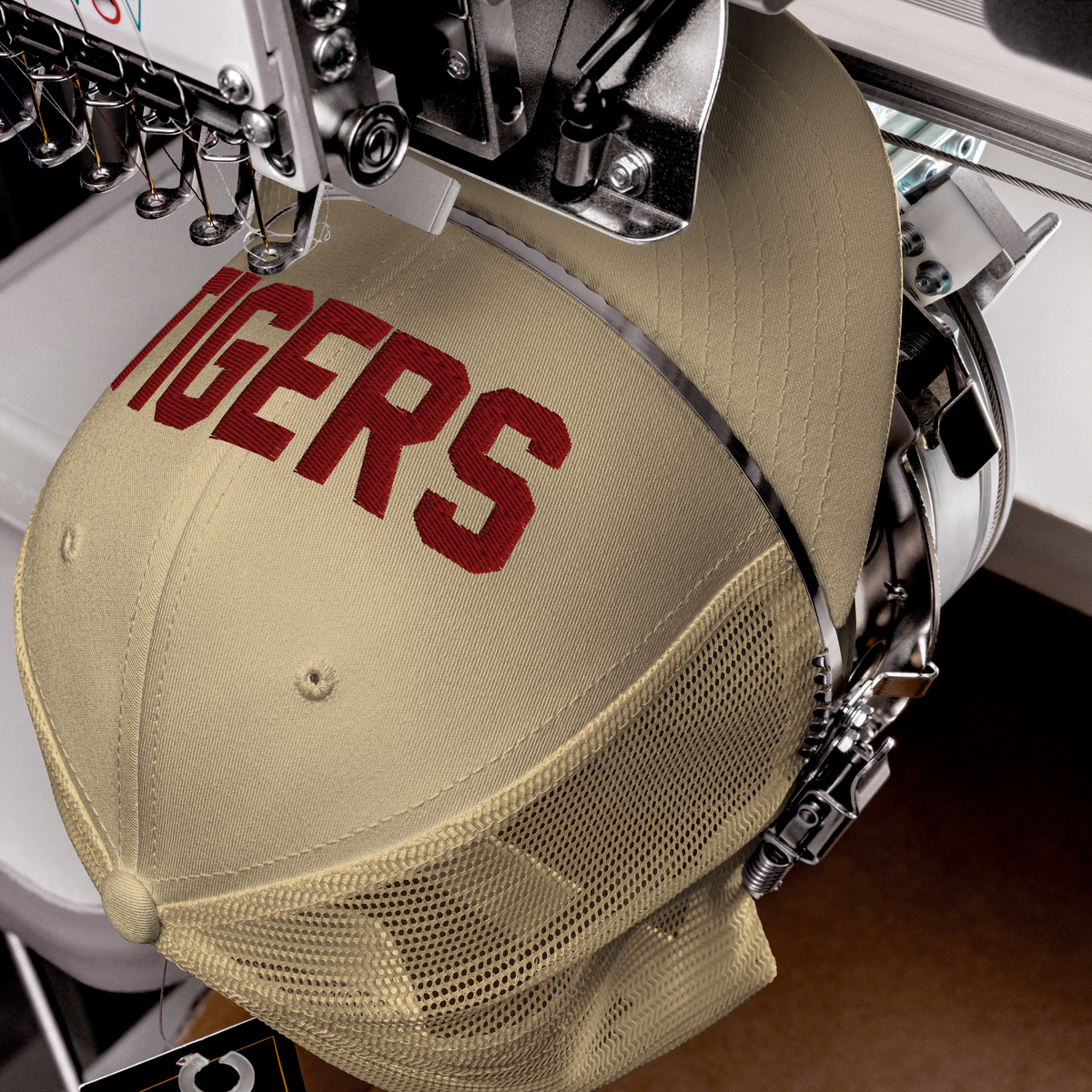 A beige baseball cap with 'TIGERS' embroidered in red on the front, shown hanging in an embroidery machine.